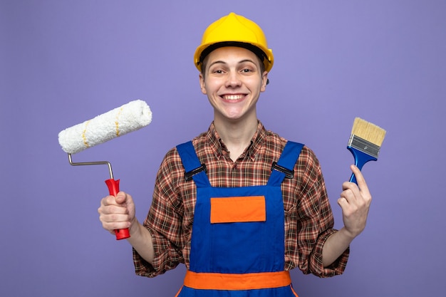 Macho joven constructor vistiendo uniforme sosteniendo pincel con cepillo de rodillo aislado en la pared púrpura