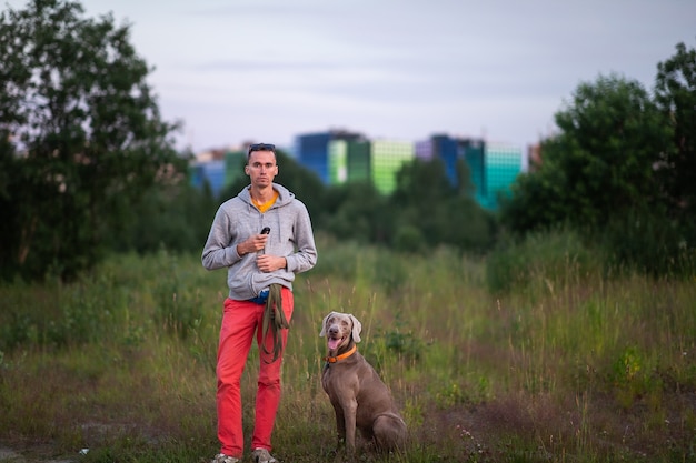 Macho joven serio en sudadera con capucha gris de pie con perro Weimaraner