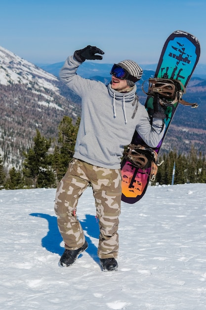 Macho joven snowboarder celebración snowboard contra el fondo de la montaña