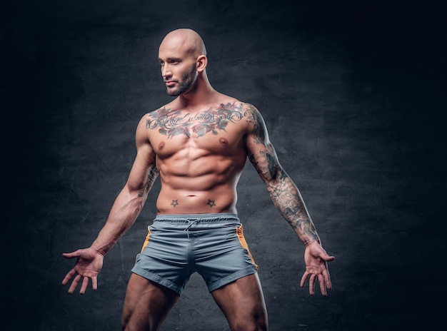 Macho musculoso sem camisa de cabeça raspada com corpo tatuado vestido com um esporte curto sobre fundo cinza.