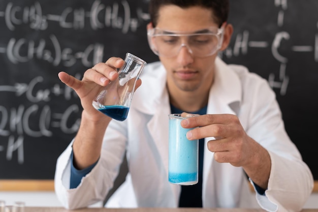 Macho profesor de química árabe mezclando líquidos haciendo experiencia en clase universitaria estudiante universitario tomando examen de química obteniendo reacción de ácidos en frasco de vidrio contra la pizarra