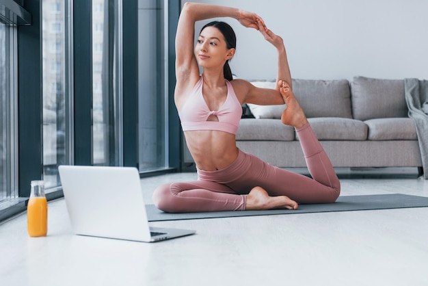 Macht Übungen auf der Matte auf dem Boden mit Laptop und einem Glas Orangensaft darauf Junge Frau mit schlanker Körperform in Sportbekleidung hat einen Fitnesstag drinnen zu Hause