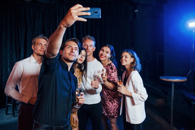 Macht Selfies. Gruppe fröhlicher Freunde, die das neue Jahr drinnen mit Getränken in den Händen feiern.