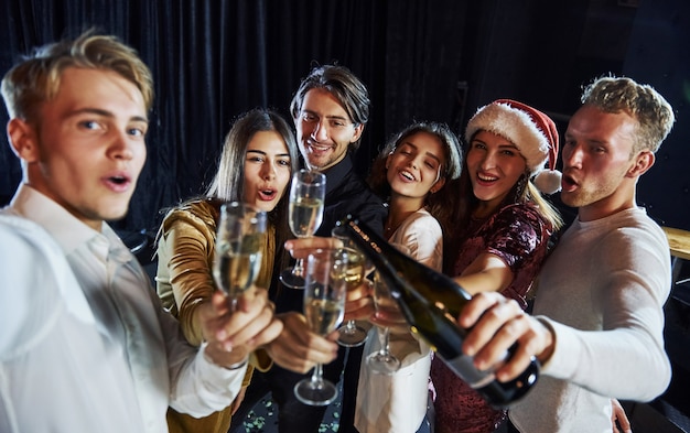 Macht Selfies. Gruppe fröhlicher Freunde, die das neue Jahr drinnen mit Getränken in den Händen feiern.