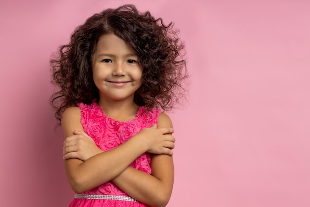 Macia menina sorridente fofa com cabelo escuro encaracolado, usando vestido, abraçando-se, olhando com expressão feliz, posando com as mãos cruzadas, isolada com espaço de cópia.