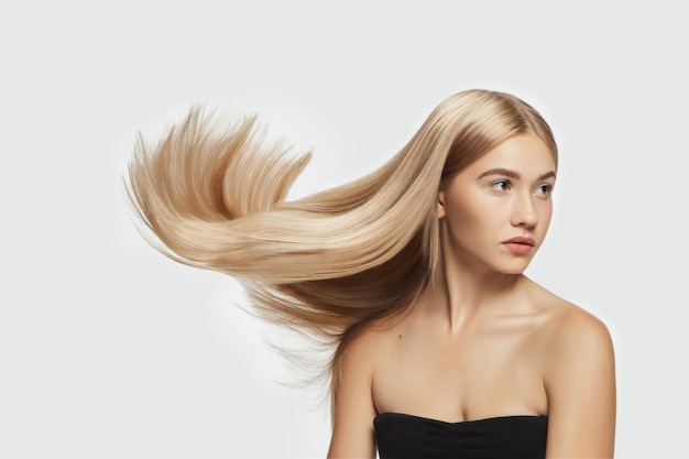 Foto macio. bela modelo com cabelo loiro longo, liso e voador em fundo branco do estúdio. jovem modelo caucasiano com pele bem cuidada e cabelos esvoaçantes. conceito de cuidados com o salão de beleza, beleza, moda.