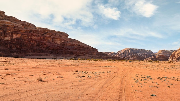 Macizos rocosos en el desierto de arena roja, cielo nublado brillante en el fondo, vehículo pequeño y camello a distancia - paisaje típico en Wadi Rum, Jordania