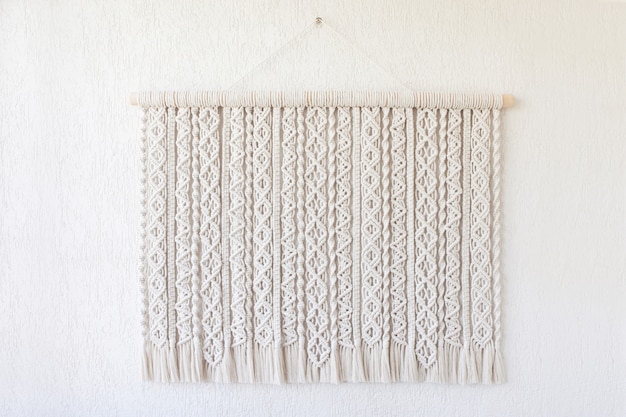 Macramé hecho a mano. Decoración de pared 100% algodón con palo de madera colgado en una pared blanca. Pasatiempo femenino. Concepto de decoración natural DIY de tejido moderno respetuoso con el medio ambiente en el interior