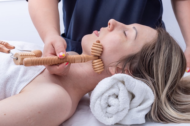 Madero-Therapie, Gesichts- und Nackenmassage, Anti-Aging-Entspannungsmassage - Die Hände des Masseurs massieren Gesicht und Hals des Mädchens mit einem Holzroller-Massagegerät, Nahaufnahme.