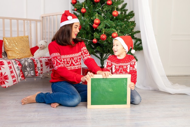 Madre abraza a un niño en suéteres rojos junto al árbol de Navidad