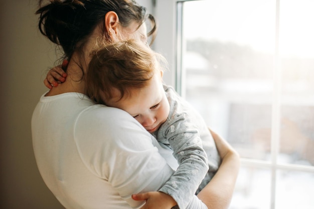 Madre abrazando a la niña mientras está de pie contra la ventana