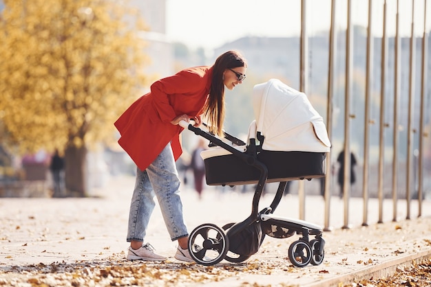 Foto madre con abrigo rojo pasea con su hijo en el cochecito en el parque en otoño.