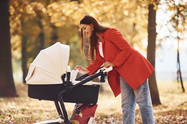 Madre con abrigo rojo pasear con su hijo en el cochecito en el parque con hermosos árboles en otoño.