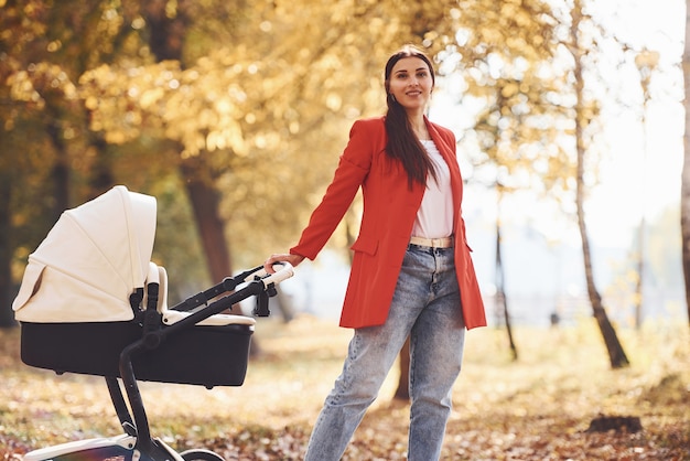 Madre con abrigo rojo pasear con su hijo en el cochecito en el parque con hermosos árboles en otoño.