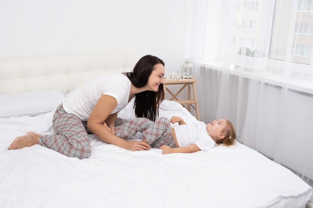 Madre adulta está haciendo cosquillas a su pequeña hija activa en la cama en casa, actividad de ocio con niños.