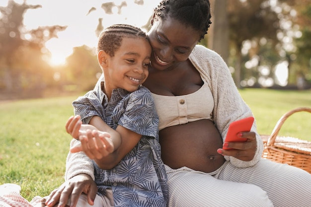 Madre africana e hijo de raza mixta mediante teléfono móvil en el parque de la ciudad - Mujer embarazada negra disfruta de un picnic con el niño al aire libre