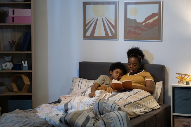 Madre afroamericana joven cuidadosa que miente con el hijo en la cama y el libro de lectura para él en la sala de guardería