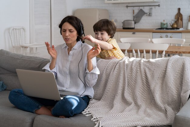 Madre agotada sentada en el sofá en casa, trabajando en la computadora portátil, niño distraído y pidiendo atención