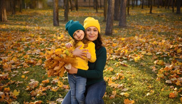 Madre alegre abrazando a su hermosa hija en un hermoso parque otoñal con arces. Mamá bonita con gorro de lana amarillo y adorable niña con sombrero verde con hojas de otoño de arce amarillo cayendo seco