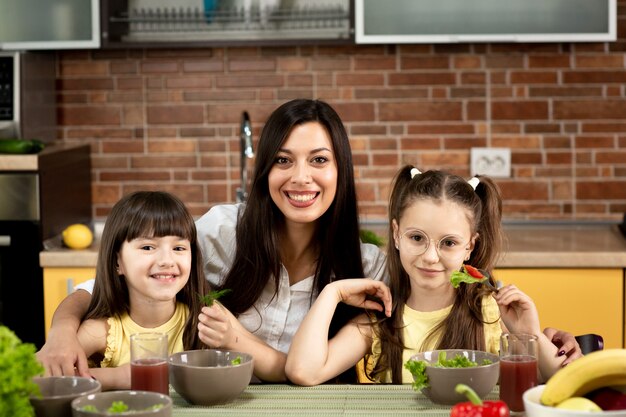 Madre alegre y dos hijas están comiendo ensalada saludable juntas en casa. El concepto de alimentación saludable, valores familiares, tiempo juntos.