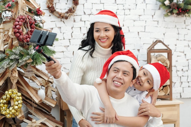 Madre alegre, padre e hijo con sombreros de santa posando para selfie familiar en el árbol de navidad