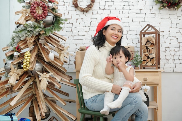 Madre alegre sentada con hija pequeña en regazos y mirando árbol de Navidad decorado