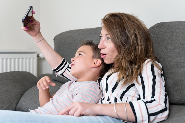 Foto madre alegre sosteniendo un teléfono inteligente haciendo selfie con su hijo pequeño mostrando el signo de la victoria, mamá y niño se sientan en el sofá usando un aparato divirtiéndose tomando autorretratos pasando tiempo juntos en casa