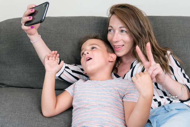 Madre alegre sosteniendo un teléfono inteligente haciendo selfie con su hijo pequeño mostrando el signo de la victoria, mamá y niño se sientan en el sofá usando un aparato divirtiéndose tomando autorretratos pasando tiempo juntos en casa