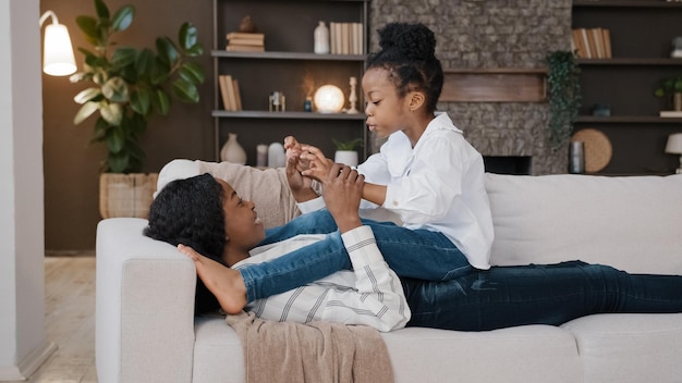 Madre amorosa afroamericana mami acostada en el sofá relajándose en la sala de estar con su pequeña hija