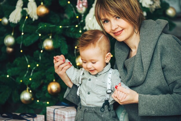 Madre amorosa con hijo en una habitación decorada para el año nuevo sonriendo juntos. Mamá feliz y bebé en el árbol de Navidad en el sillón. La primera Navidad del niño.