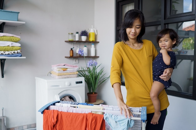 Madre asiática ama de casa secando y colgando ropa en el lavadero en casa mientras lleva a su bebé
