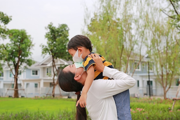 Madre asiática cargando a su hijo con mascarilla protectora y besándose en un jardín público durante el brote de coronavirus y gripe