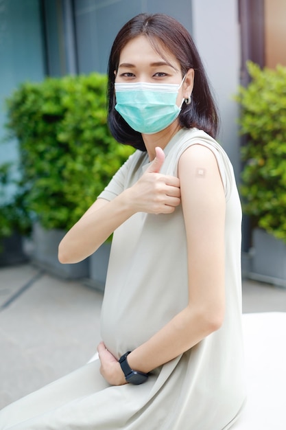 Una madre asiática embarazada con un vestido largo lleva una máscara y el pulgar hacia arriba después de vacunarse en el Servicio Nacional de Salud.