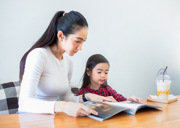 Una madre asiática está enseñando a su hija a leer un libro durante el descanso del semestre en la mesa de estar y tomando leche fría en la mesa en casa. Conceptos educativos y actividades de la familia.