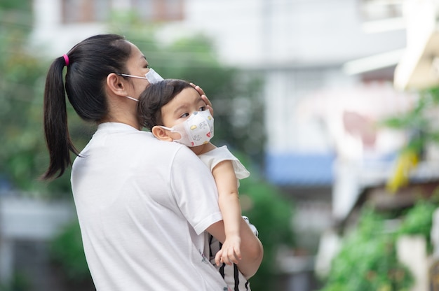 Foto madre asiática con mascarilla blanca sosteniendo a su pequeña niña pequeña que es su hija usa mascarilla de bebé, mirando hacia afuera, concepto covid-19