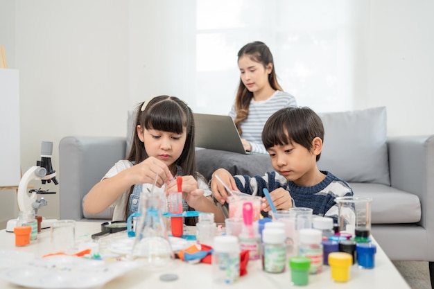 Madre asiática mira a su hija e hijo haciendo experimentos químicos y pruebas en la sala de estar en casa