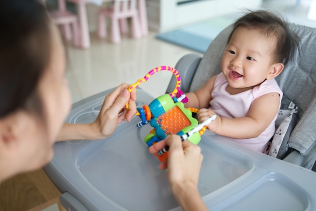 Madre asiática que juega el juguete con su bebé que se sienta en la silla dinning en casa.