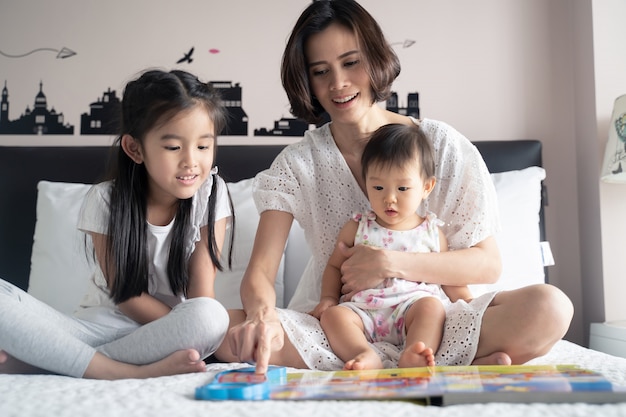 Madre asiática que lee el libro de música a sus hijas que se sientan en la cama.