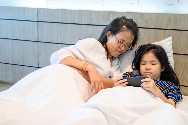 Madre asiática y su hija están usando un teléfono inteligente y sonriendo en la cama en su casa