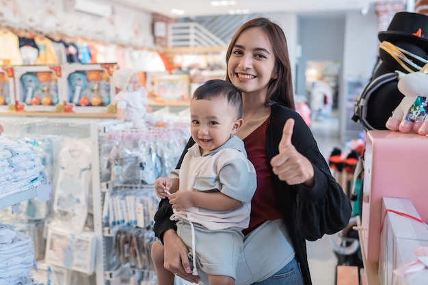 Madre asiática con su niño pequeño de compras en la tienda del bebé con el pulgar hacia arriba