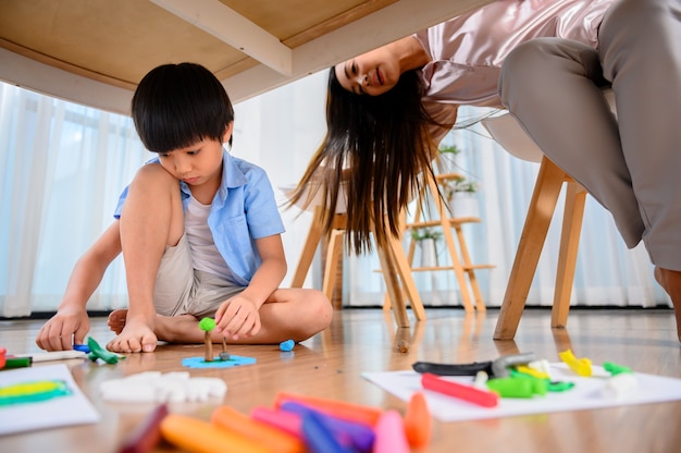 Madre asiática trabaja en casa junto con su hijo. Mamá trabajando en línea y plastilina para niños debajo de la mesa. Estilo de vida de la mujer y actividad familiar.