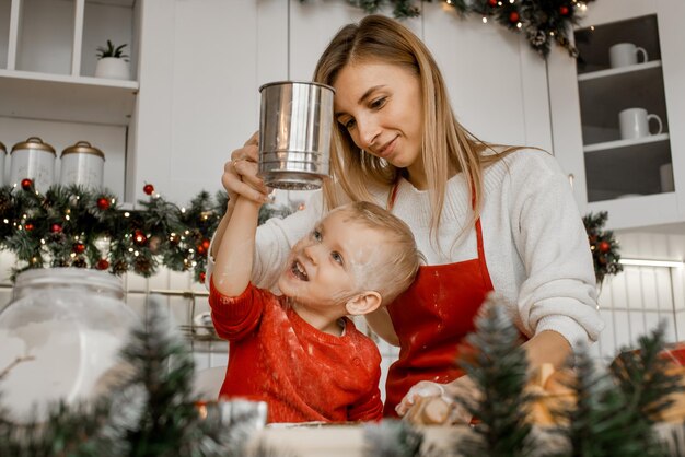 Madre atractiva joven ayuda a su pequeño hijo encantador en un suéter rojo tamizar la harina a través de un colador en la cocina de Navidad. El muchacho está mirando el tamiz y riendo.