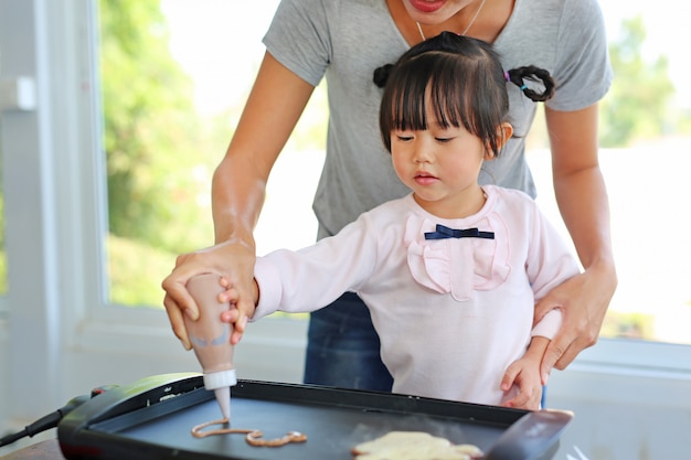 Madre ayudando a su hija a verter la harina mezclada en la sartén para hacer tortitas