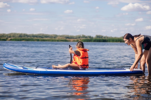 Madre ayudando a su pequeña hija a cenar con remo en las manos en el lago con juncos verdes y árboles en el fondo con chaleco salvavidas Vacaciones activas Inculcación del amor por los deportes desde la infancia