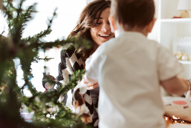 Madre y bebé decorando y jugando alrededor del árbol de Navidad