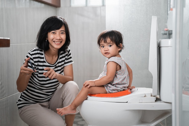 La madre y el bebé sonrientes miran a la cámara mientras miran videos en teléfonos móviles en el inodoro en el baño