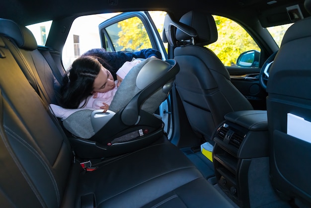 Madre besando a su bebé en el asiento de bebé en el asiento trasero del automóvil con interior negro, visto desde el otro lado del vehículo con espacio de copia