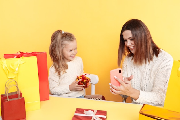 Madre bonita con hija pequeña sentada en el escritorio con regalos y usando el teléfono para elegir regalos en línea.