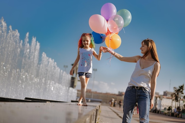 Madre caminando con su hija de la mano a lo largo de la fuente de la ciudad Niña descalza sosteniendo globos