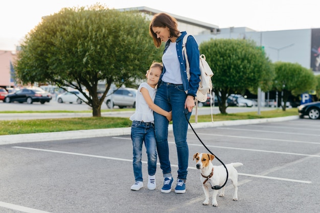 La madre cariñosa abraza a la pequeña hija, da un paseo al aire libre junto con su mascota favorita